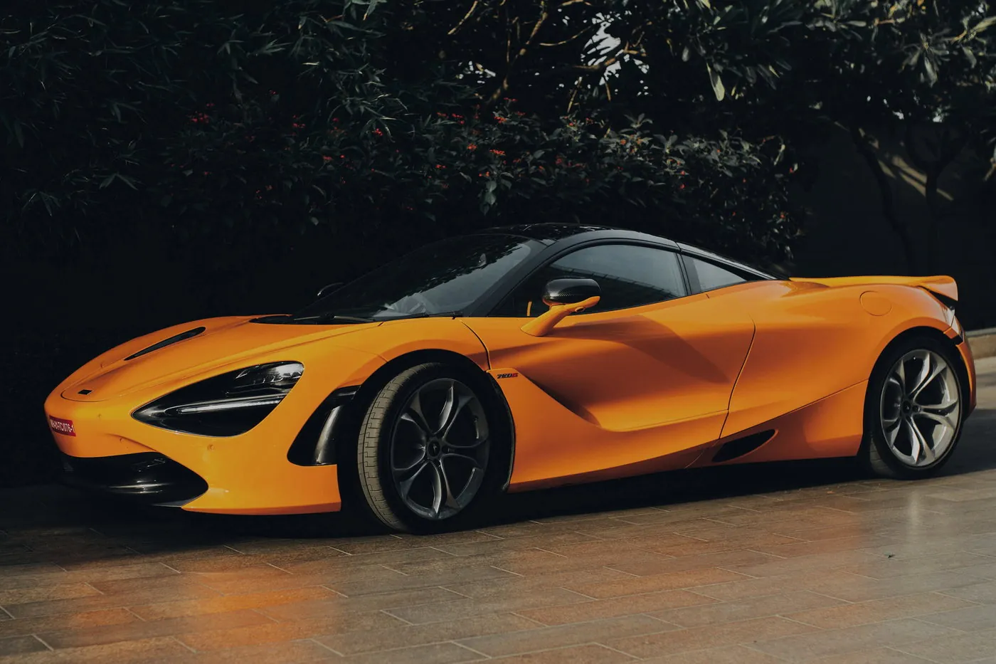 McLaren 720S arancione papaya, tre quarti anteriore, davanti a una siepe scura al tramonto